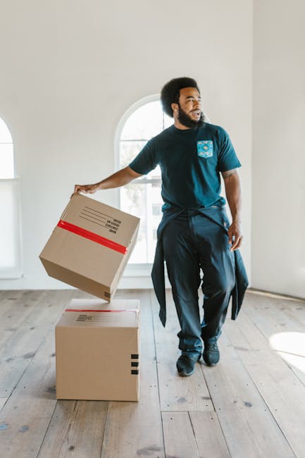A man in a black shirt and blue jeans is pushing a heavy-duty trolley loaded with large cardboard boxes and packing materials, including plastic wrap, along an outdoor pavement area under an elevated