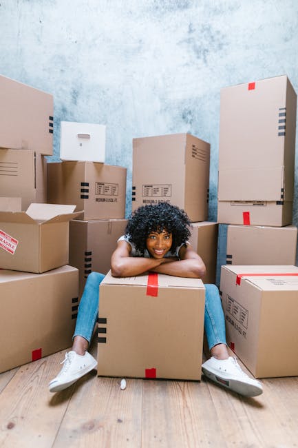 A man with curly hair and dark skin seated on the wooden floor of a room, surrounded by several cardboard boxes with red packing tape, some stacked next to him and others on the floor. He is wearing a